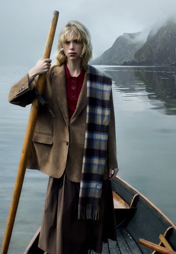 Brauner Wildleder-Blazer über einem roten Hemd mit Knöpfen. Umschlungen mit einem blauen und beigen karierte Schal. Dunkelbrauer Rock; hält ein Holzrudern.