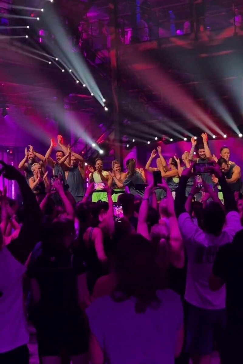 Groep artiesten op het podium in felle kleding met een mix van groen en blauw, temidden van een menigte in een schemerige ruimte met paarse verlichting.