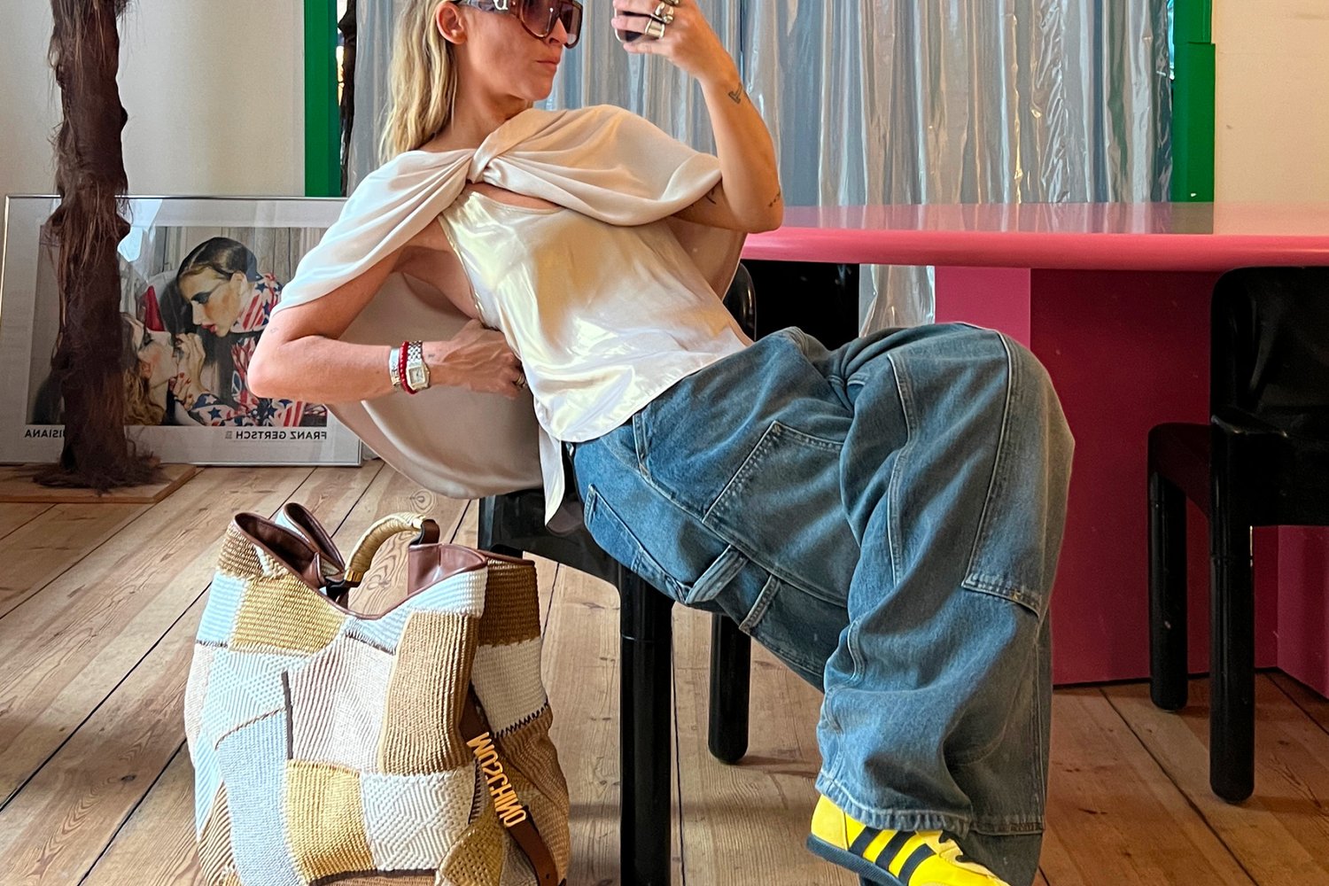 Mujer con gafas de sol y top de satén reclinada en una silla negra junto a un bolso de patchwork, una mesa colorida y una obra de arte enmarcada sobre el suelo de madera.
