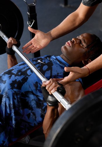 Una persona che indossa una canotta atletica blu e nera a motivi sta effettuando una panca con un bilanciere, tenendo il bilanciere con entrambe le mani, in un ambiente di palestra.