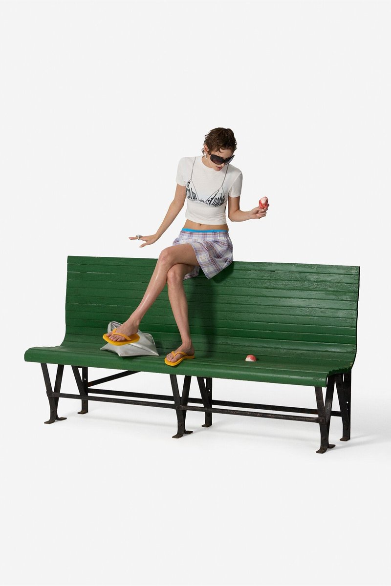 Young woman in sunglasses, white crop top, and plaid skirt sits on green bench eating an ice cream cone with one foot resting on a bag.