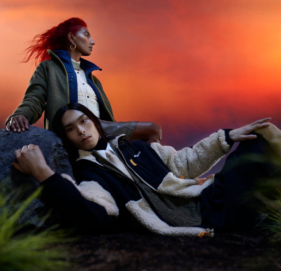 Two individuals pose against a dramatic sunset. One stands confidently, while the other reclines on a rock, both in stylish outerwear.