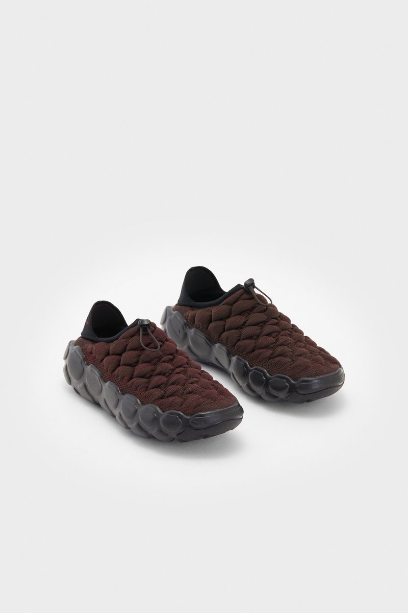 Zapatos de slip-on tejidos en un rojo oscuro con suelas negras, que presentan un patrón texturizado y un cordón elástico para un ajuste seguro, sobre un fondo blanco.