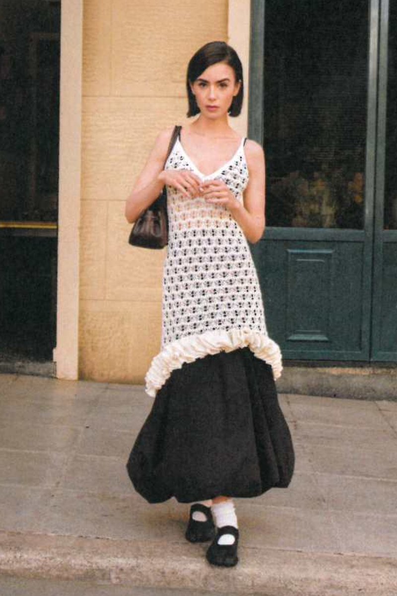 Femme portant une robe blanche en crochet sans manches sur une longue jupe noire, chaussures Mary Jane noires avec chaussettes blanches, debout devant l'entrée du bâtiment.