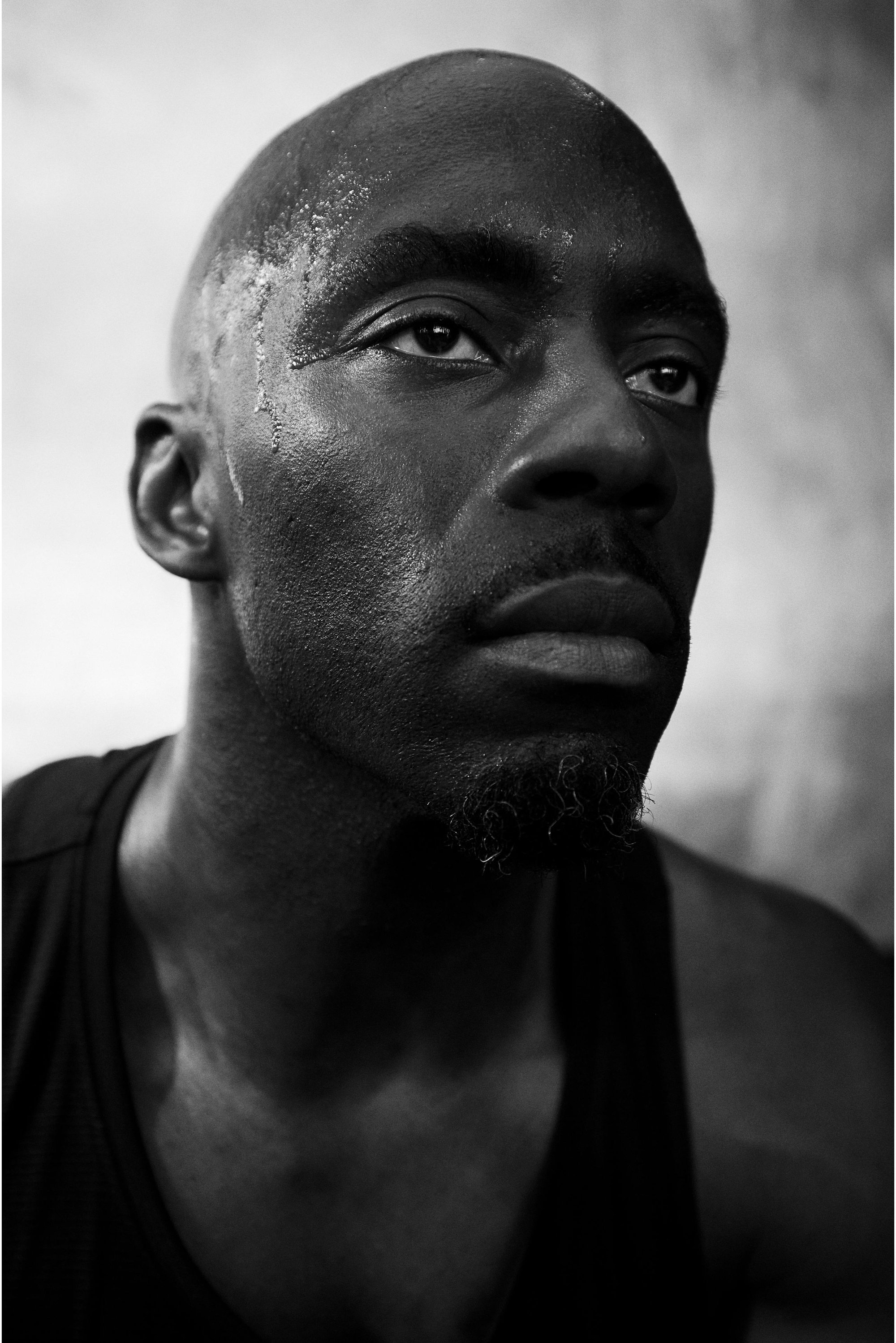 Detailný záber na muža, ktorý má pot drenážou a sústredený výraz, oblečeného v tielku, na rozmazanom neutrálnom pozadí.