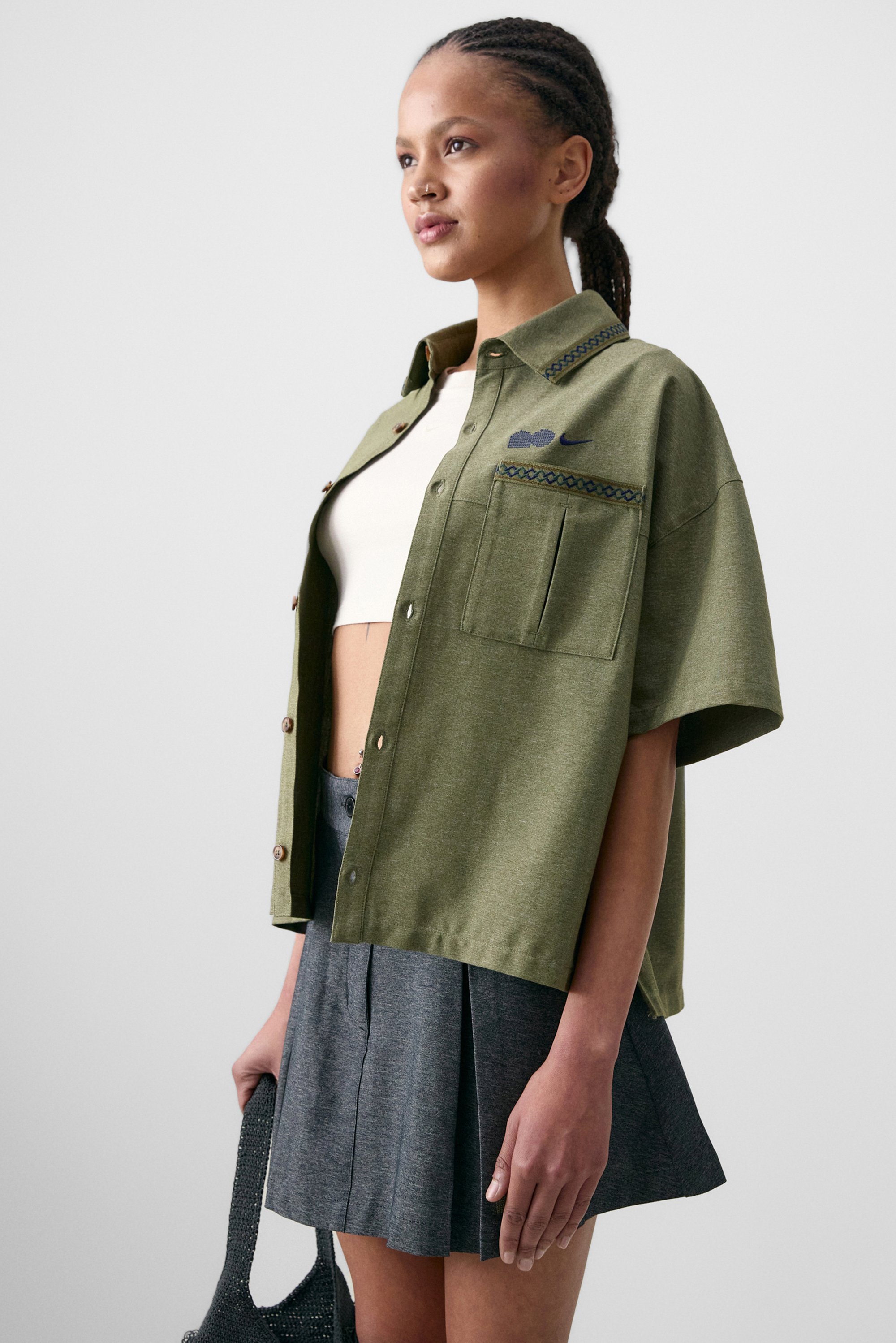 Model wearing an olive green oversized shirt over a white crop top, paired with a gray pleated skirt, holding a black bag. Neutral background.