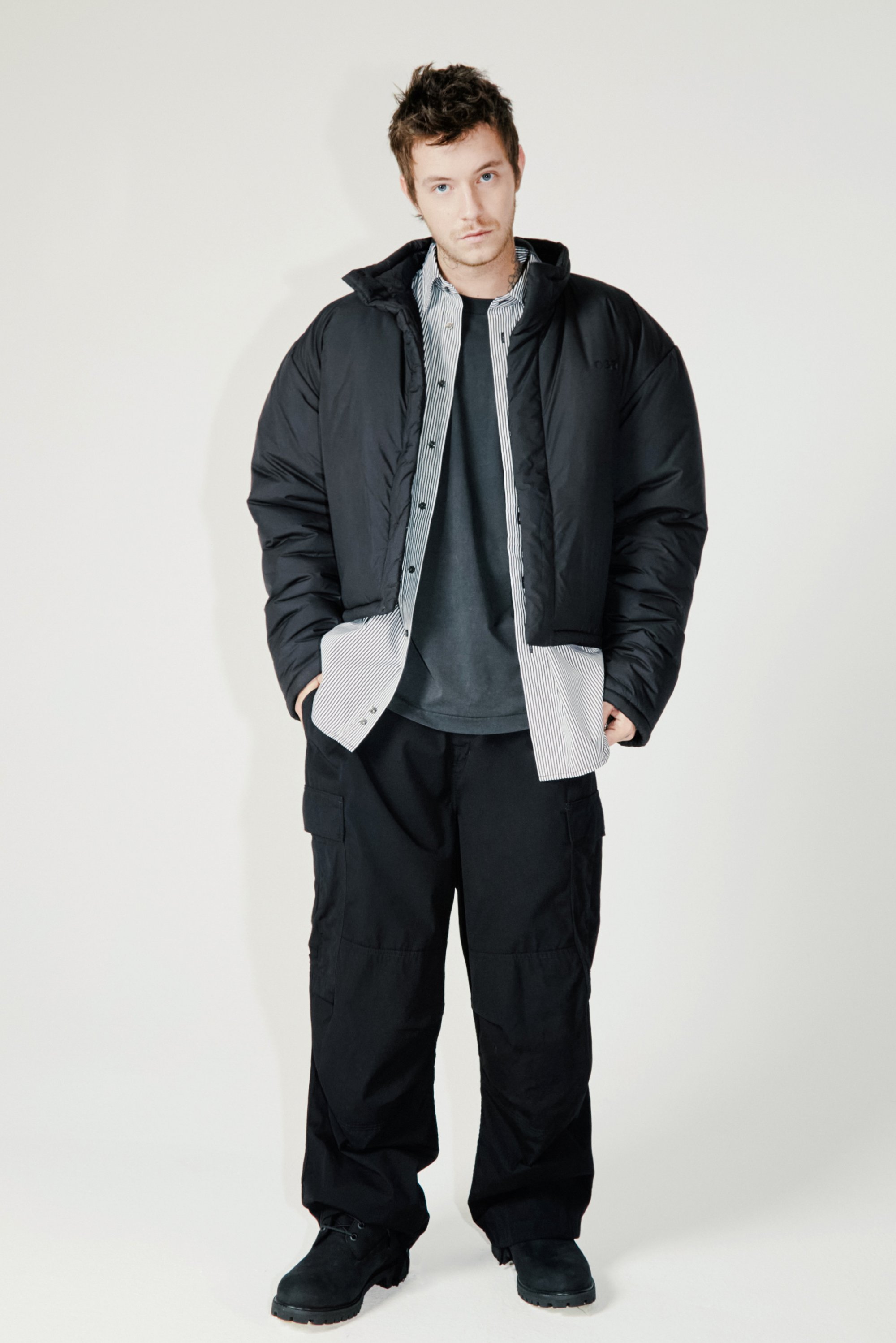 Chaqueta de plumas negra con capucha, llevada sobre una camisa gris y una camisa de botones a rayas. Pantalones cargo negros y botas a juego completan el look.