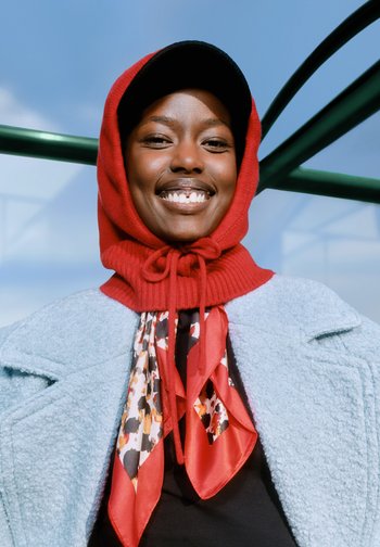 Rød strikhætte med snøre, lagt over en blå tekstureret frakke. Sort top under med et rødt og mønstret tørklæde om halsen.