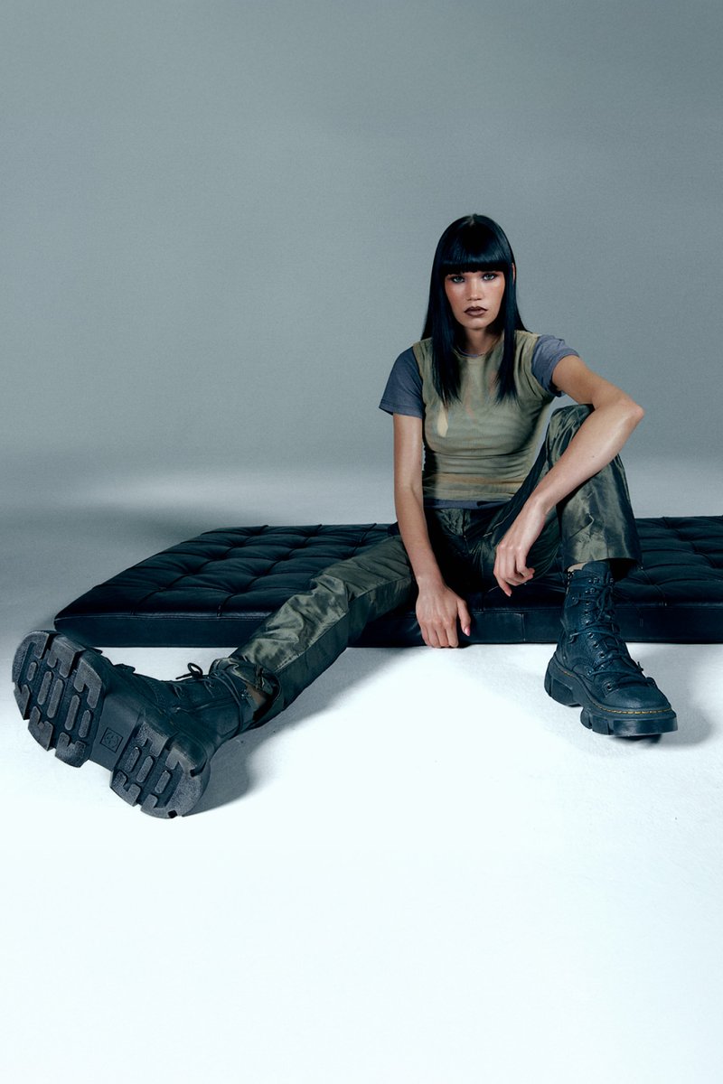 Una modelo con cabello largo y negro está sentada sobre una superficie acolchada negra, vistiendo una blusa verde y gris, pantalones cargo oliva y botas de combate negras.