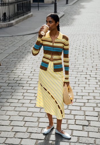 Femme en cardigan rayé et jupe jaune boit dans un gobelet en papier tout en tenant un sac tressé dans une rue pavée.