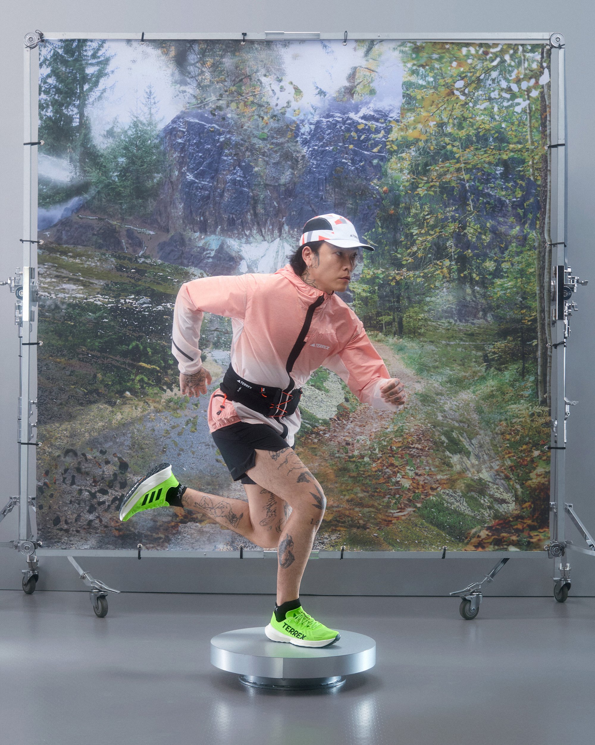 Una persona corre su un palco rotante, indossando un giubbotto rosa, pantaloni neri e scarpe verde brillante, con uno sfondo naturale.