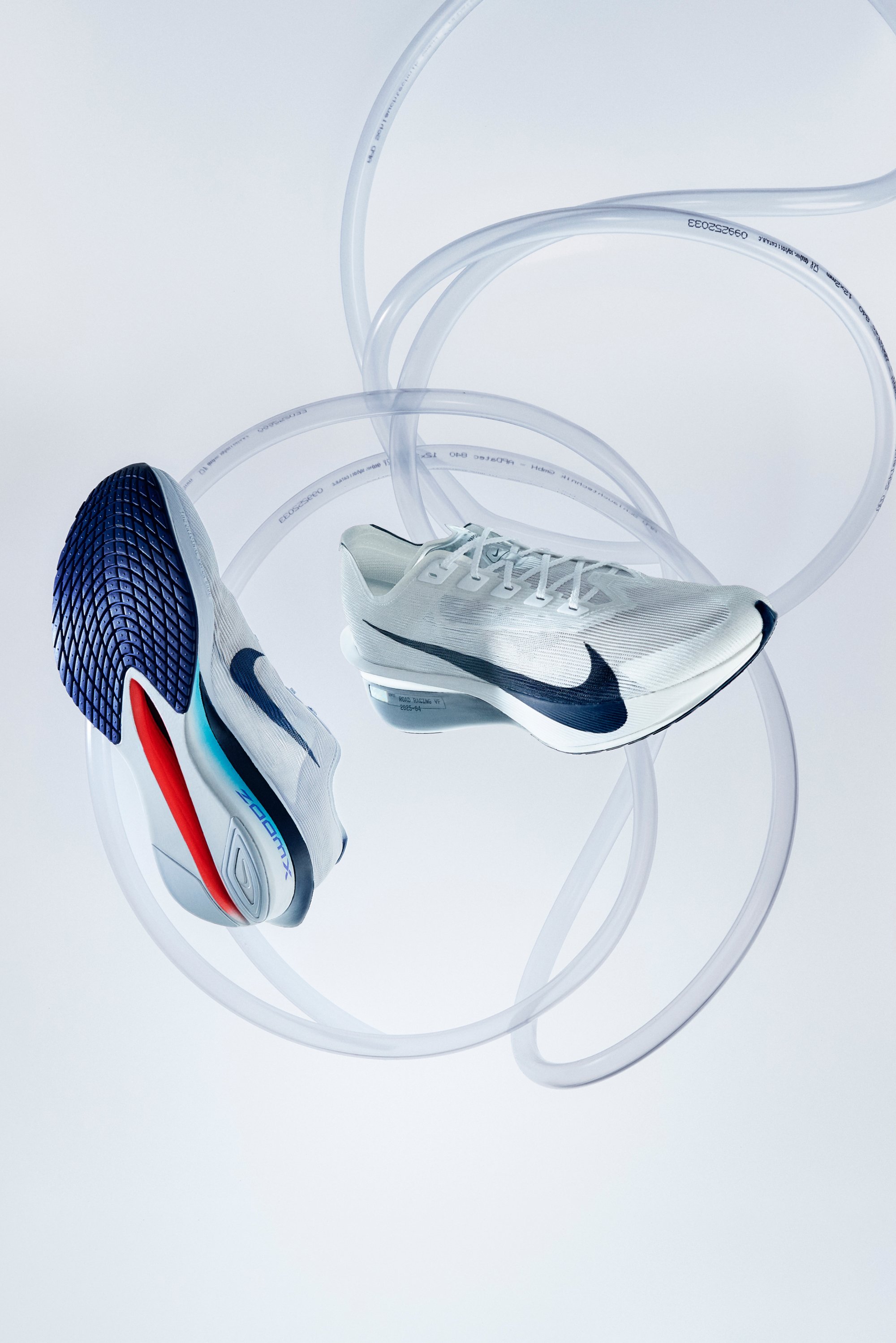 Weiße Laufschuhe mit einem Obermaterial aus Mesh, blauen und roten Akzenten sowie einer strukturierten schwarzen Sohle, die in klarem Tubus vor einem hellen Hintergrund aufgehängt sind.