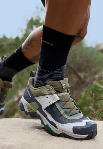 Person wearing black socks and green, white, and grey trail running shoes stepping on a rocky surface outdoors.