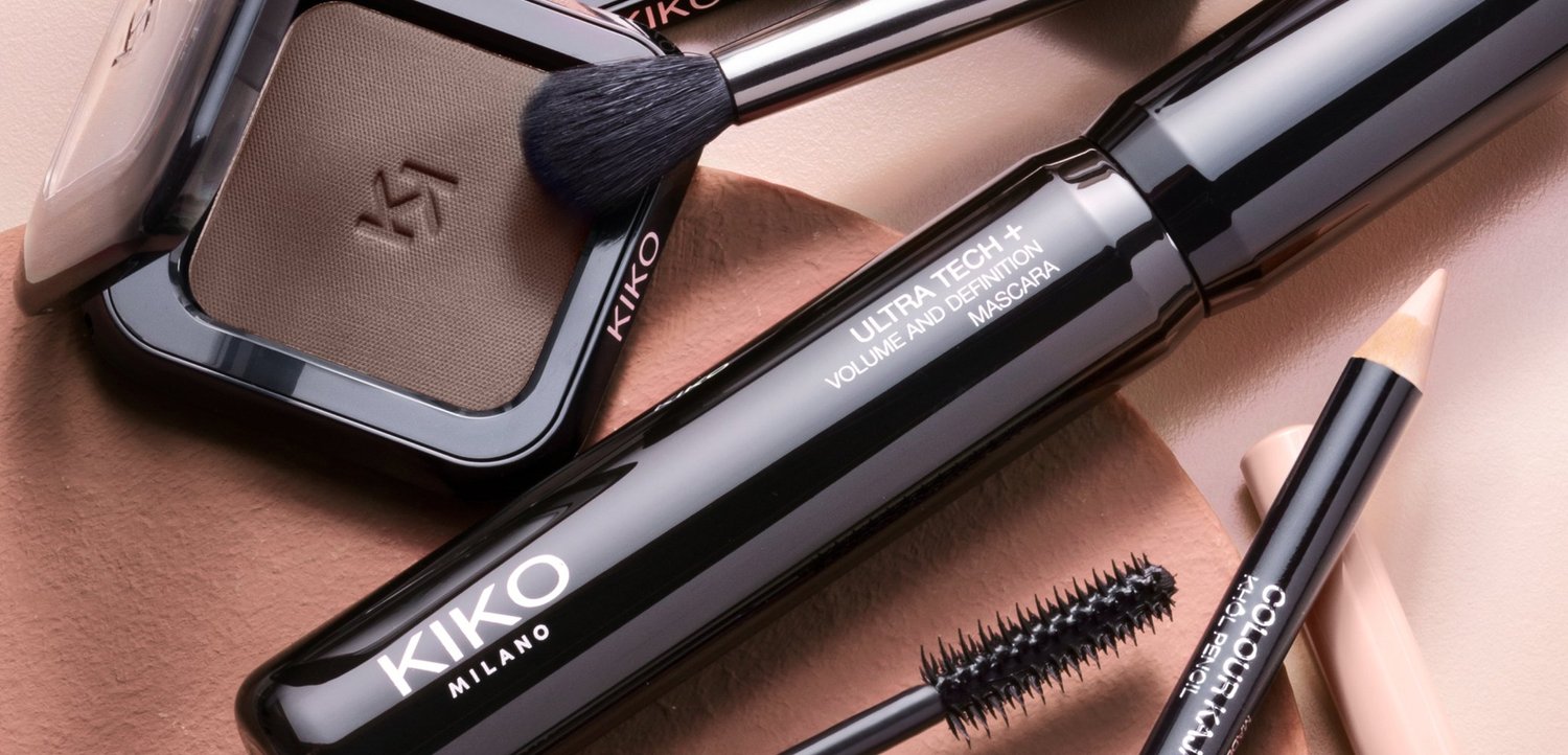 Kiko Milano make-up artikelen, waaronder bruine oogschaduw, volumemascara, een schuine kwast en een beige eyelinerpotlood, op een bruine ondergrond.