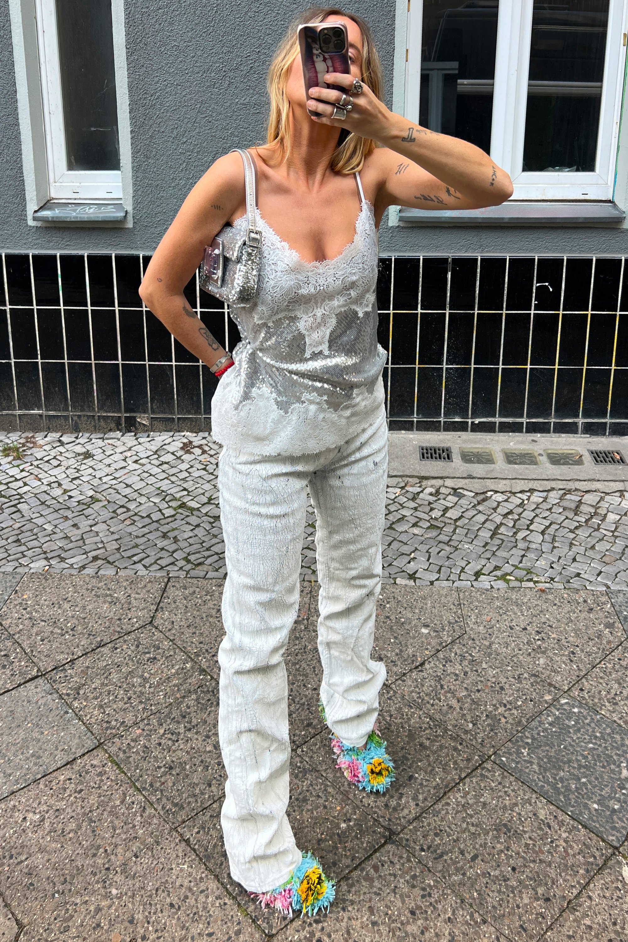 Donna in un top di pizzo bianco e paillettes con pantaloni abbinati, pantofole colorate e pelose, borsa a spalla argento, mentre tiene il telefono per un selfie all'aperto.