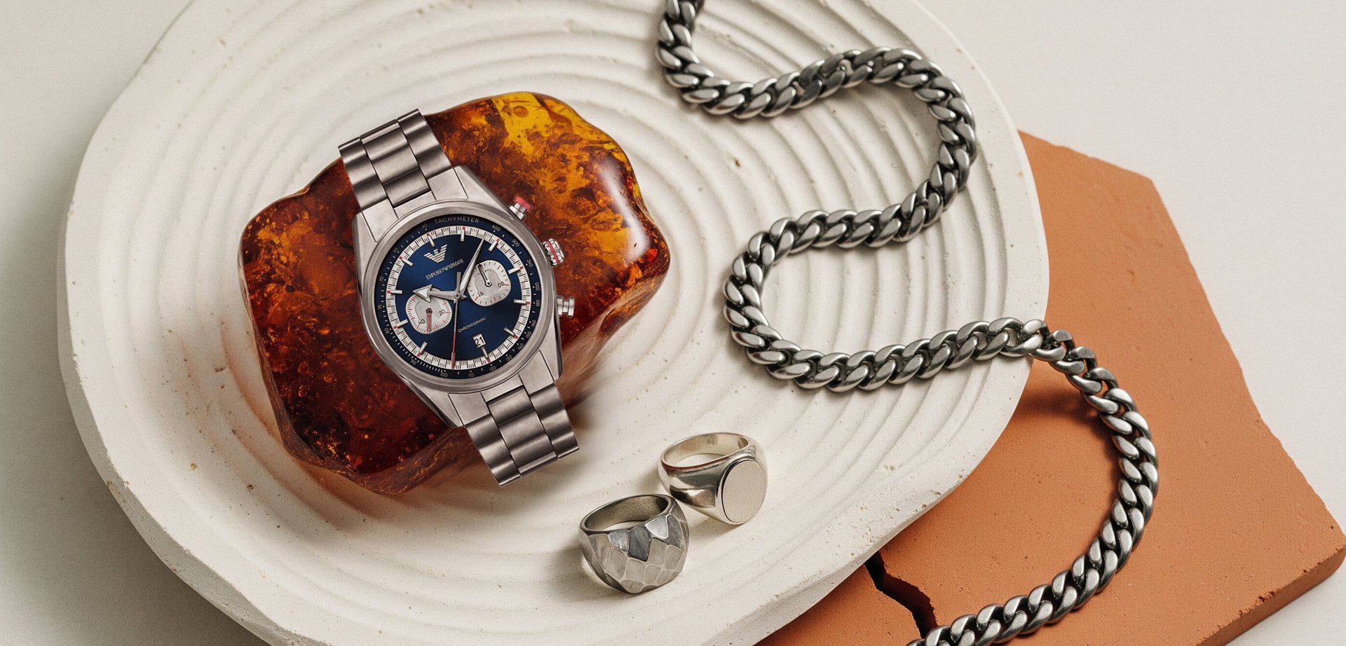 Silver chronograph watch with a blue face resting on a brown amber stone. It features two dials, a date display, and nearby are silver chains and rings.