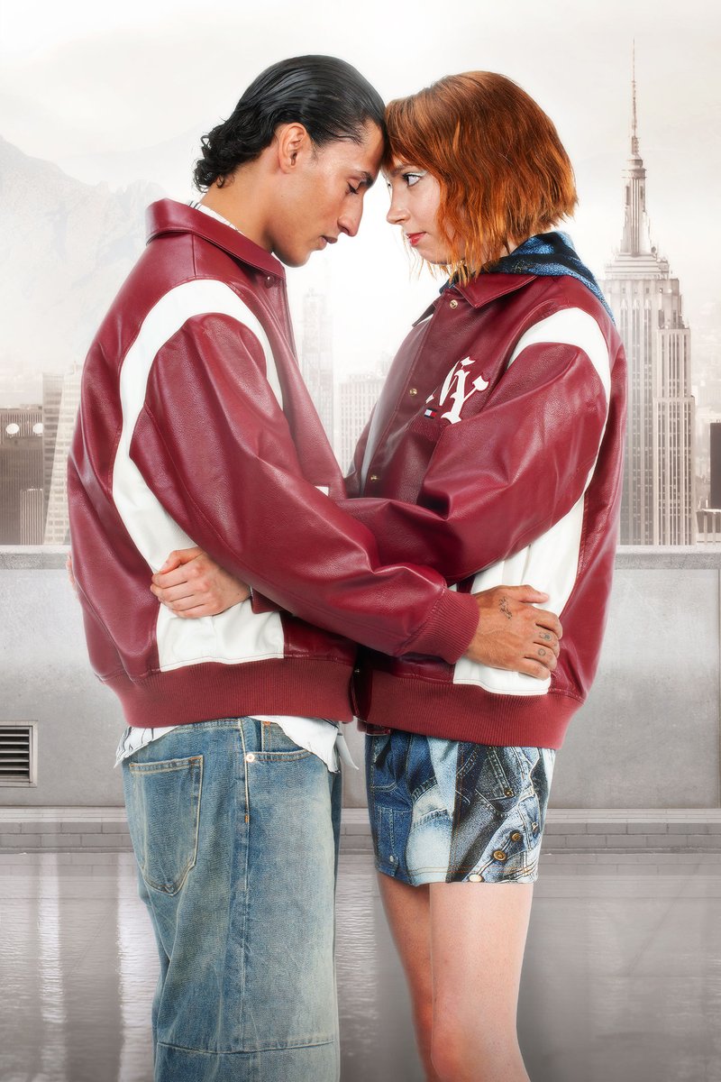 A man and woman in matching burgundy jackets embrace on a rooftop, with a city skyline and skyscrapers in the background.