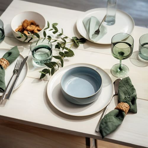 Stół udekorowany jasnym drewnianym blatem, okrągłymi białymi i niebieskimi talerzami, zielonymi serwetkami z naturalnym sznurkiem oraz zielonymi naczyniami szklanymi.