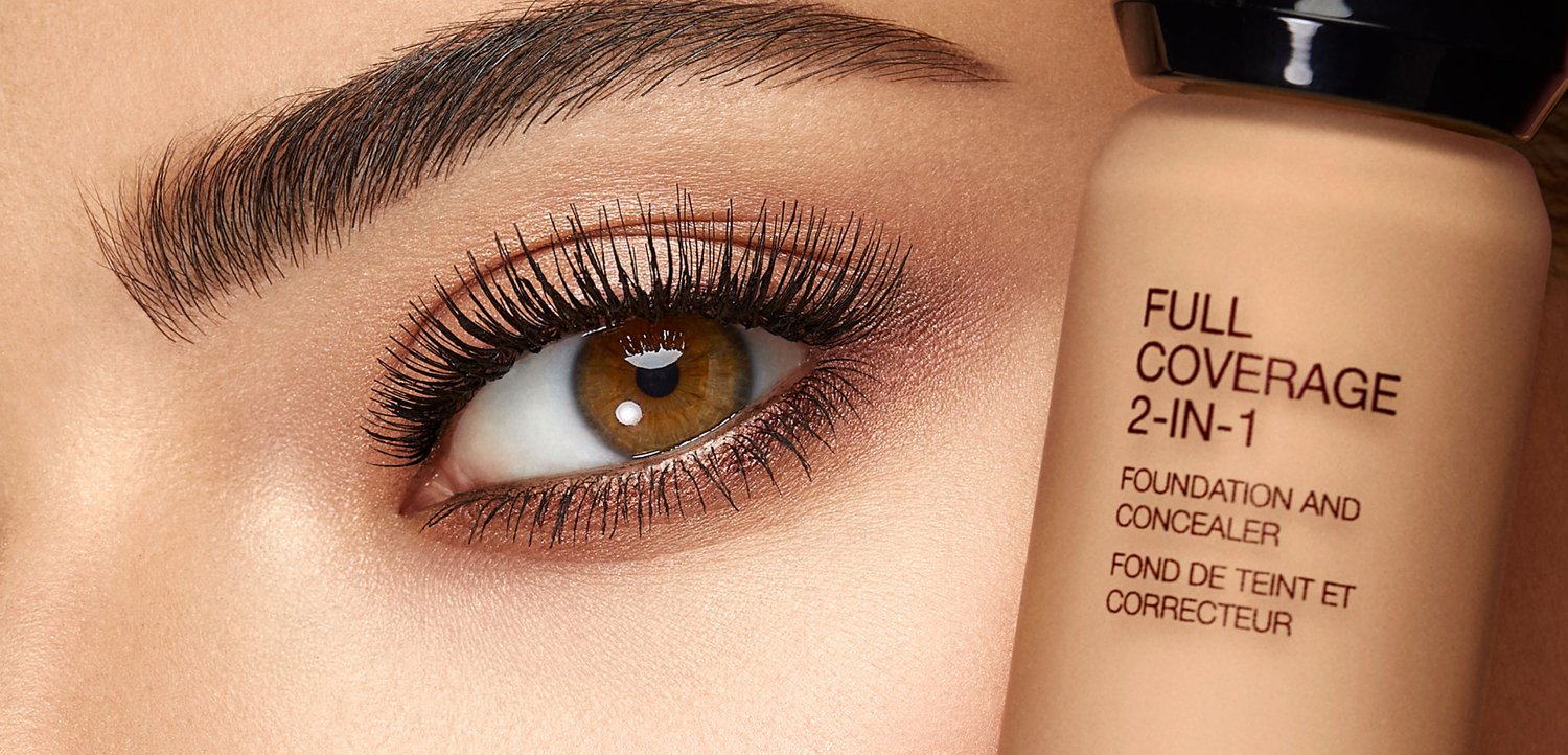 Gros plan d'un œil marron avec de longs cils et un sourcil façonné à côté d'une bouteille étiquetée fond de teint et correcteur 2 en 1 couvrance totale.