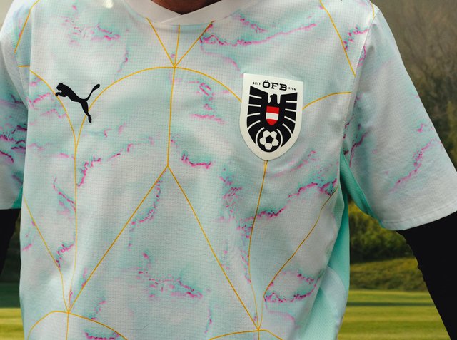 Biała i pastelowo niebieska koszulka piłkarska z różowymi akcentami, żółtymi geometrycznymi liniami, czarnym logo Puma i herbem Austriackiego Związku Piłki Nożnej na piersi.