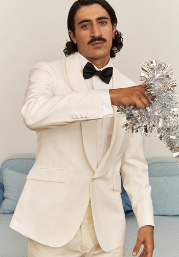 Hvit smokjakke med satengslag, parret med hvit skjorte og svart sløyfe. Holder sølvfrynse, står på en lyseblå sofa.