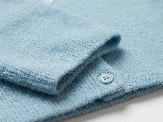 Primo piano di un maglione lavorato a maglia azzurro chiaro piegato con texture visibile e due bottoni rotondi su una superficie bianca.
