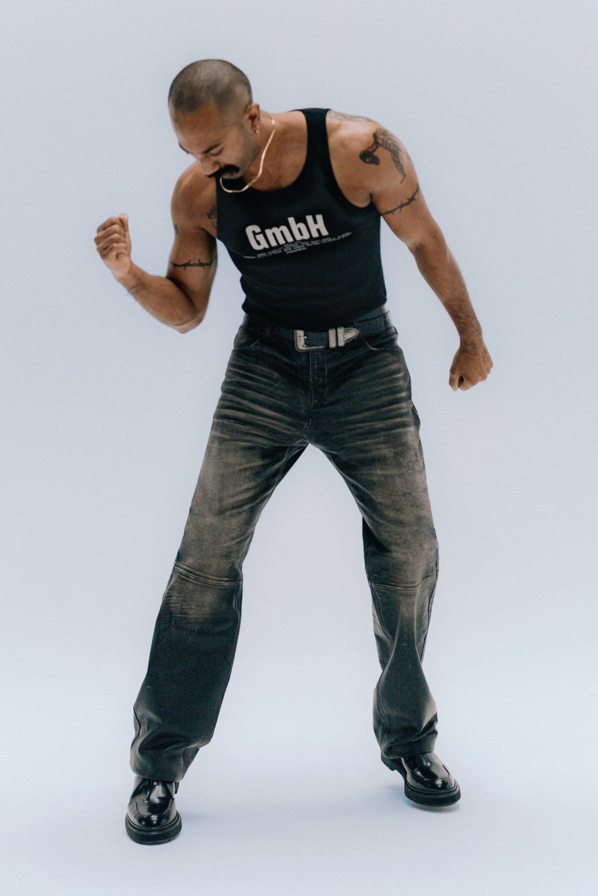 Una persona con la cabeza rapada y tatuajes lleva una camiseta sin mangas negra y jeans desgastados, posando de forma confiada en una pose de baile contra un fondo claro.