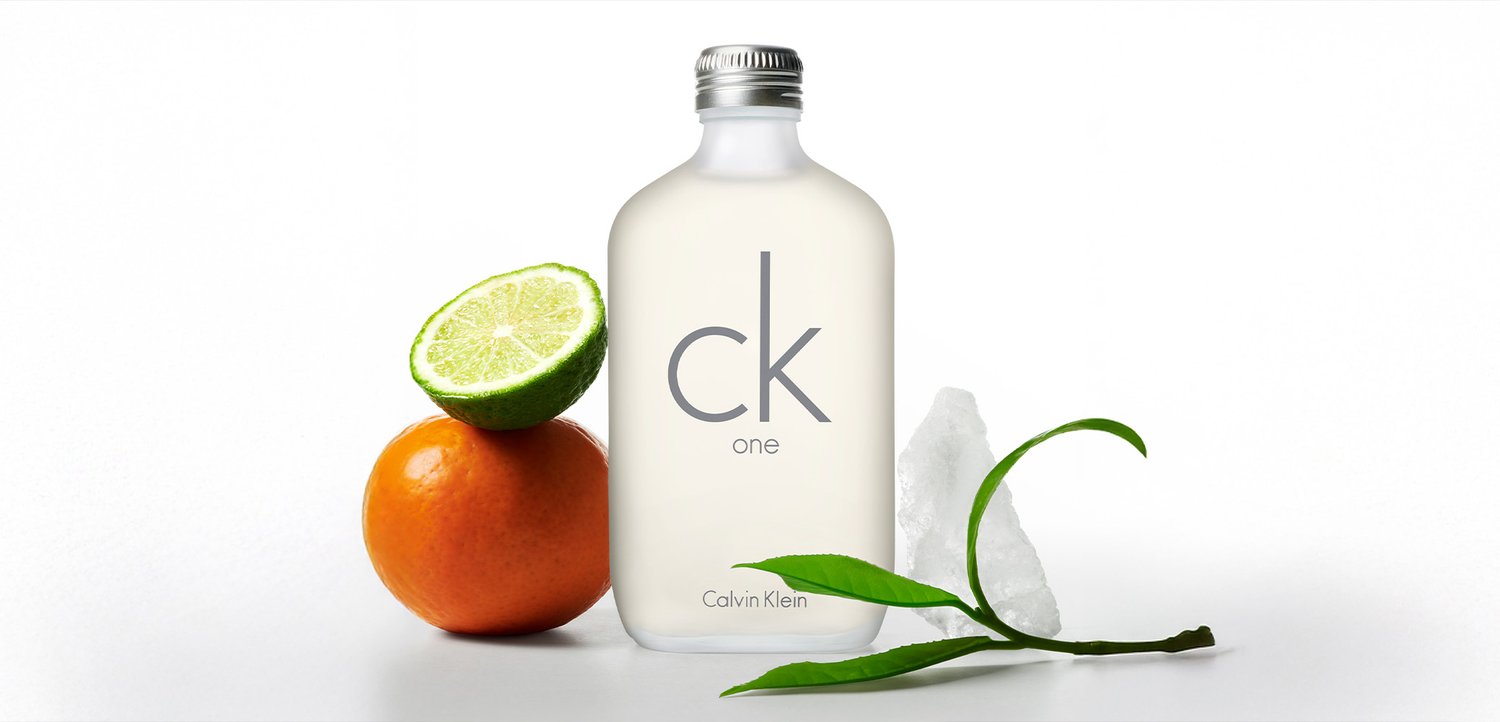 Bottiglia di profumo Calvin Klein CK One con una metà di lime, arancia, cubetti di ghiaccio e foglie verdi su uno sfondo bianco.