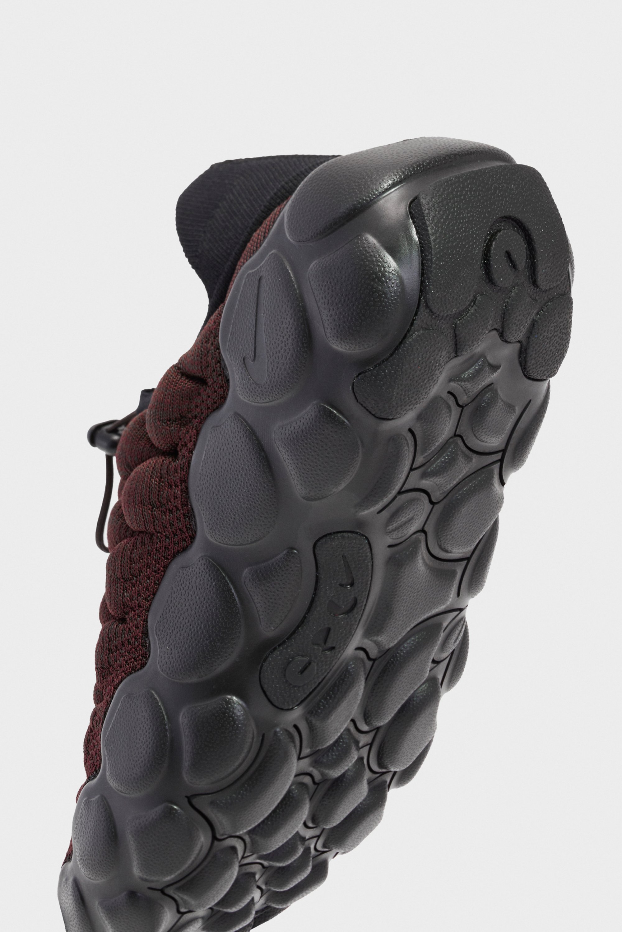 Close-up view of a shoe sole featuring a textured black rubber pattern and maroon knit upper. The shape is modern and athletic.
