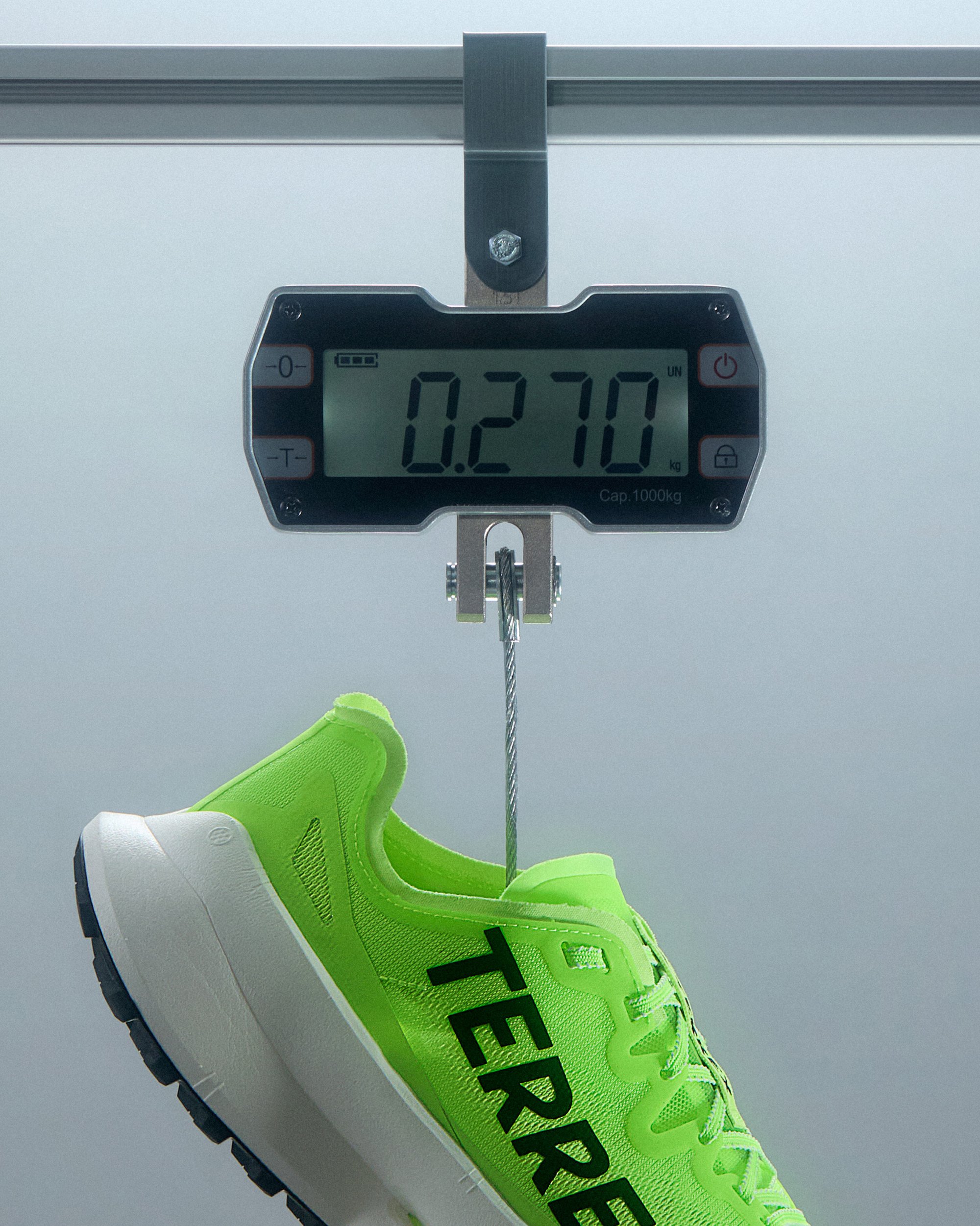 Una sneaker verde brillante etichettata "TERRE" pende da una bilancia che mostra un peso di 0,270 kg su uno sfondo grigio.