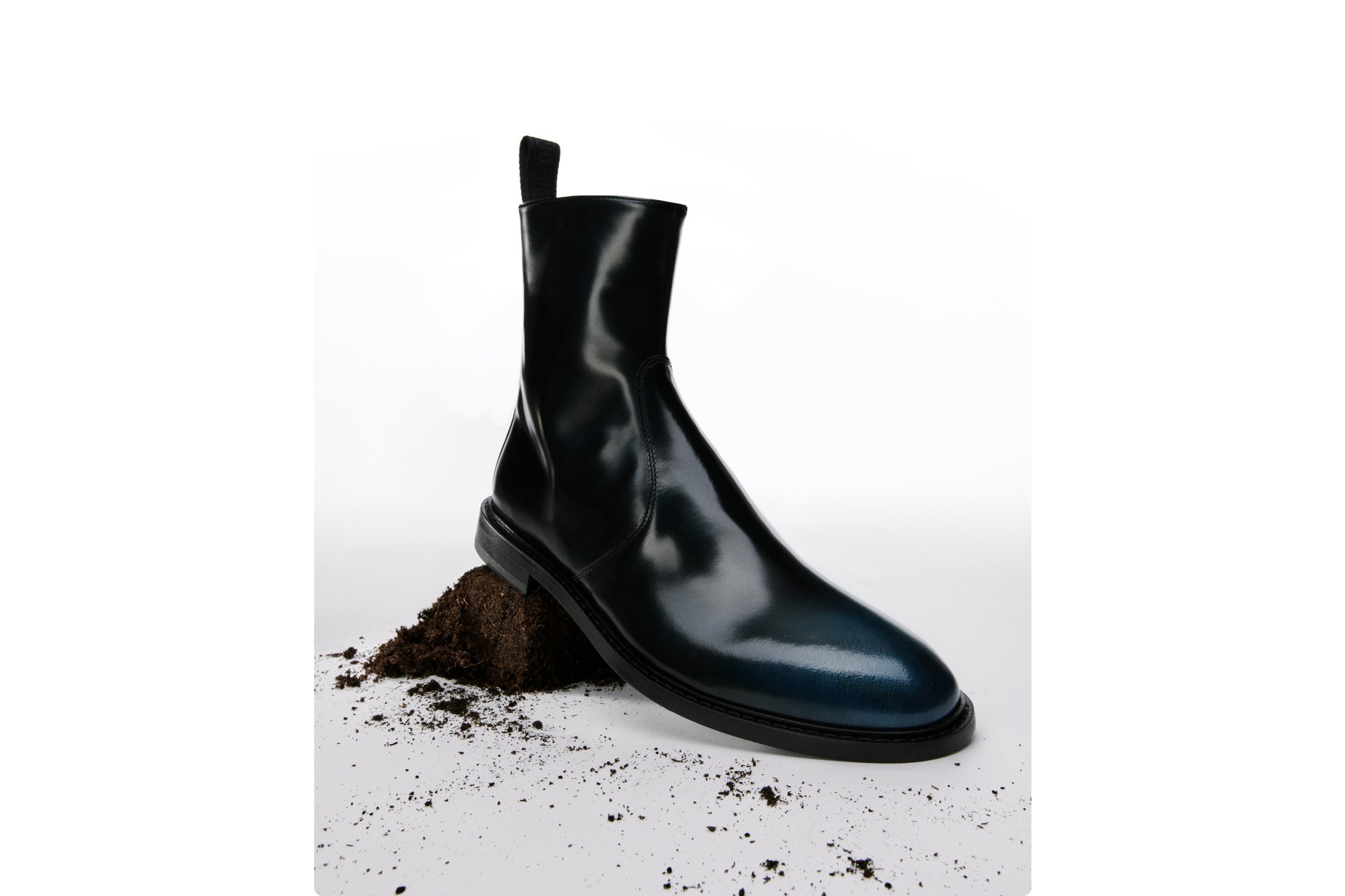 Glanzende zwarte enkelboot rustend op een hoop donkere aarde, waarbij het slanke ontwerp en de glanzende afwerking tegen een witte achtergrond worden benadrukt.