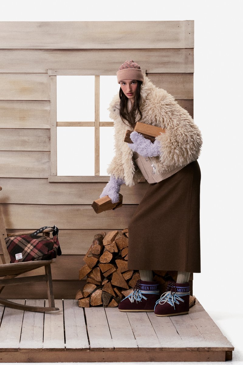 Woman wearing winter clothes stacking firewood inside a wooden cabin with a tartan bag on a rocking chair nearby.