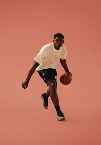 Un homme dribble un ballon de basket tout en courant, portant une chemise blanche et un short noir, sur un fond rose.
