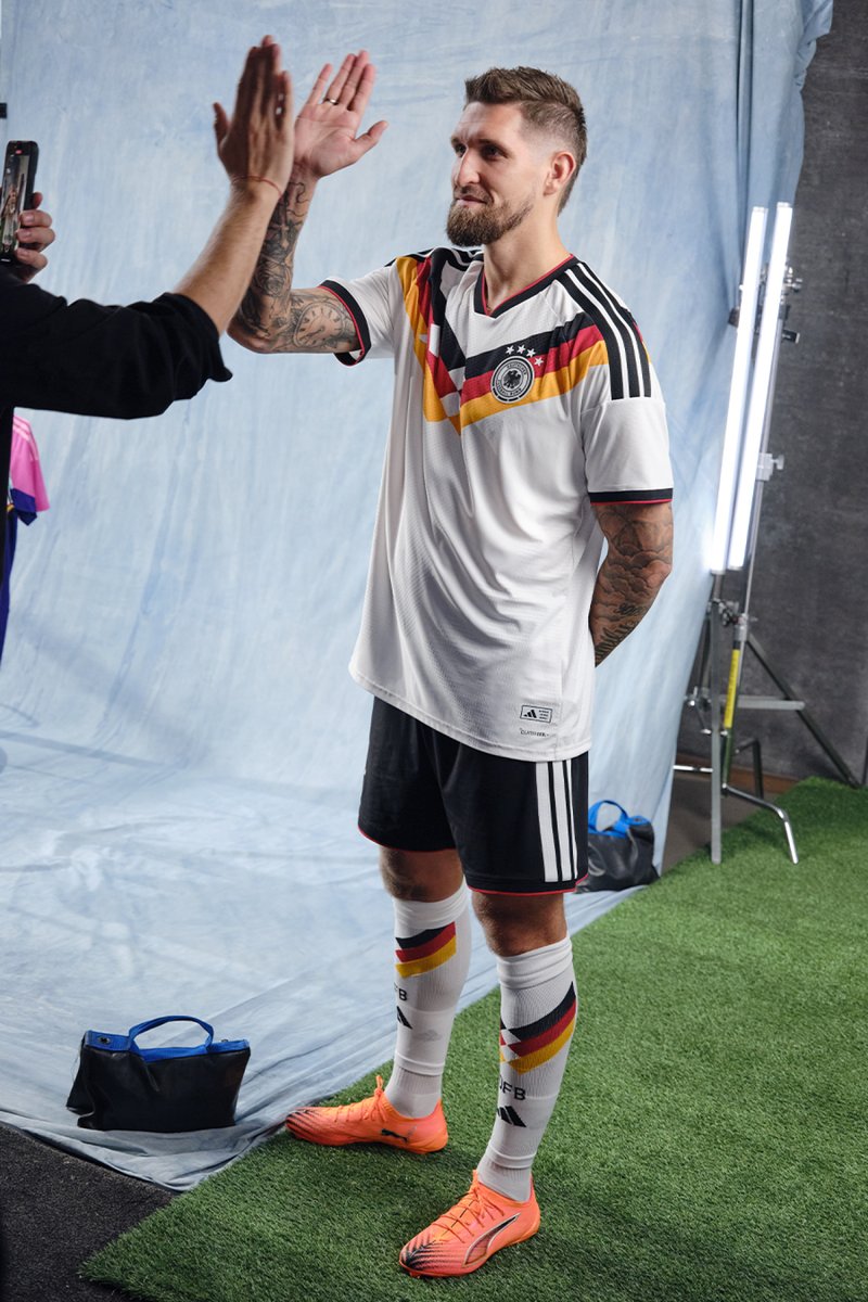 White football shirt with black, red, and yellow trim; black shorts; orange football boots; player wearing socks in national colours; standing on artificial turf.