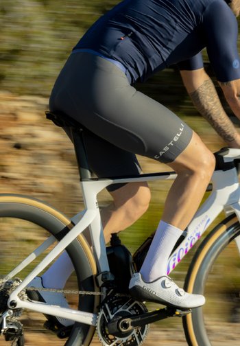 Cycliste portant un maillot bleu marine et un short gris roule sur un vélo de route blanc avec des pneus couleur sable sur un sentier extérieur flou.
