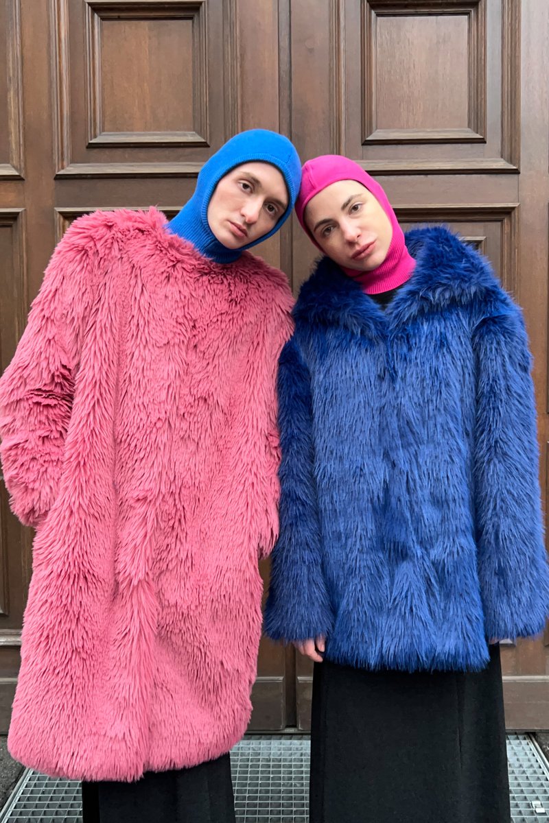Two models pose closely, wearing oversized, fuzzy coats—one pink, one blue—against a wooden backdrop. Both wear matching colorful hats.