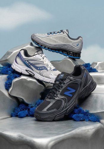 Tres zapatillas deportivas en gris, blanco y negro con detalles azules, exhibidas sobre rocas plateadas con musgo azul bajo una luz suave.