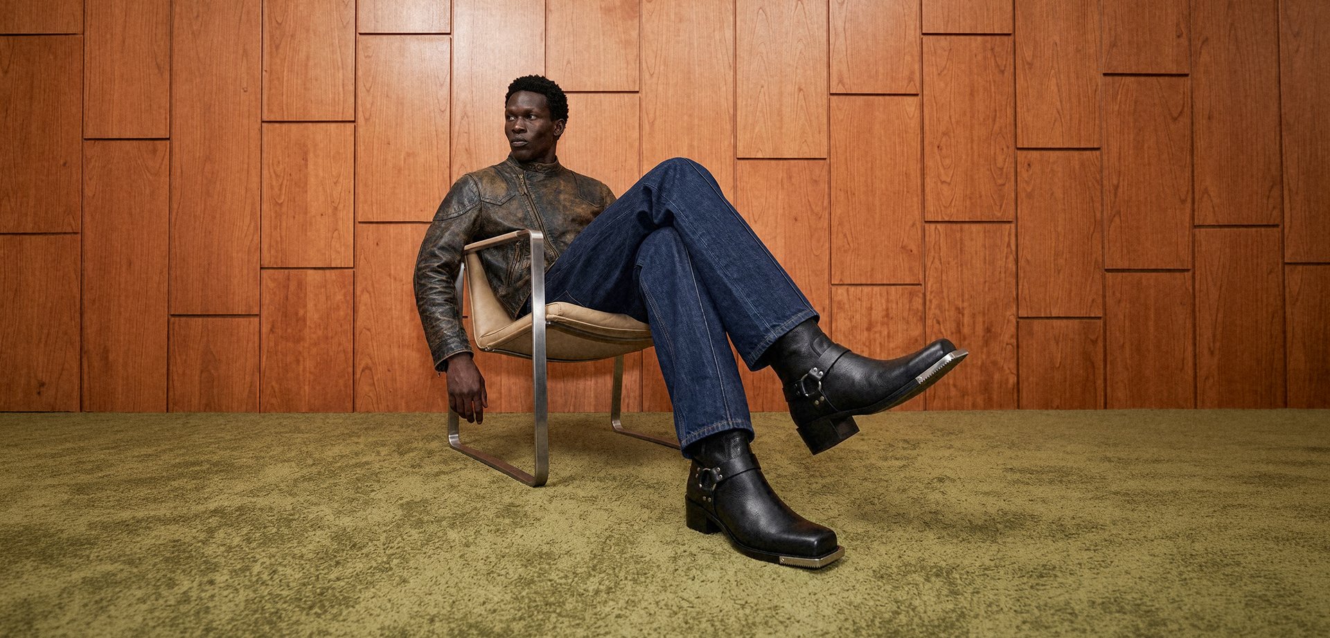 Homme portant une veste en cuir marron et un jean assis sur une chaise moderne devant un mur en bois avec un tapis vert.
