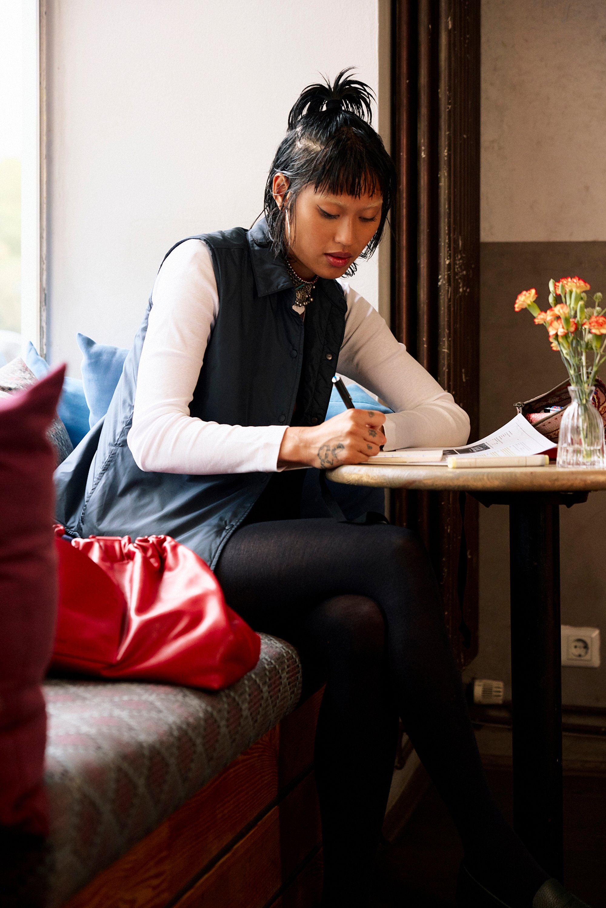 Black padded vest over a white long-sleeved shirt, seated beside a red handbag on a patterned bench, writing on paper at a table.