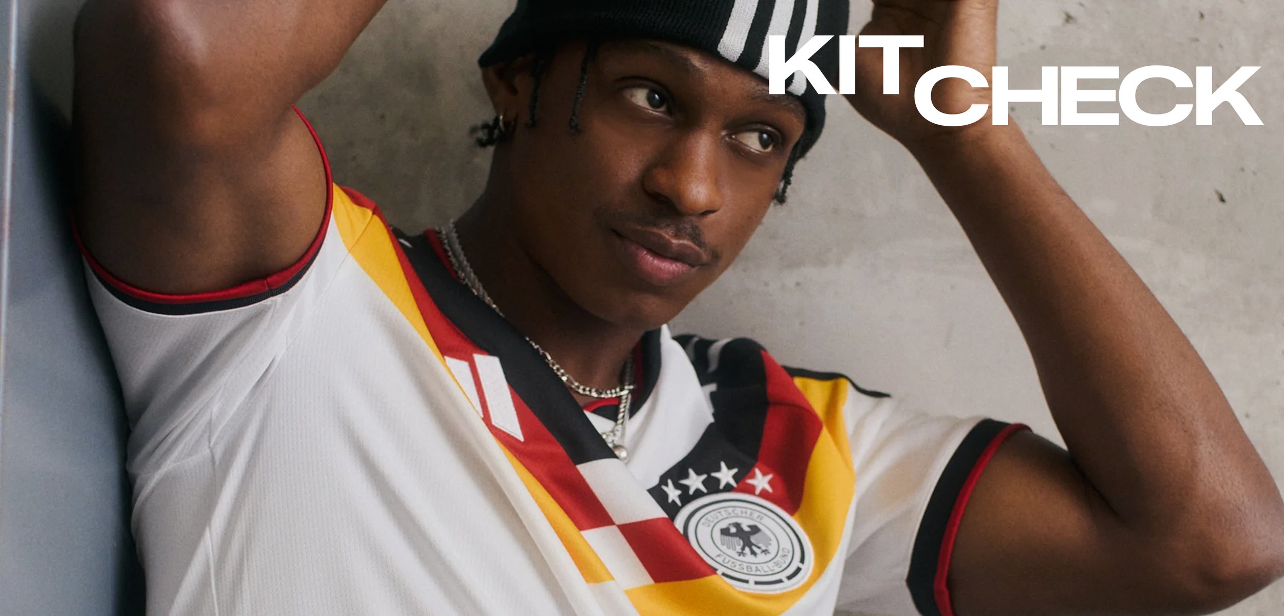 Junger Mann richtet schwarz-weiss gestreiften Hut, trägt ein mehrfarbiges Sporttrikot mit Emblem und Text "KIT CHECK" auf grauem Hintergrund.