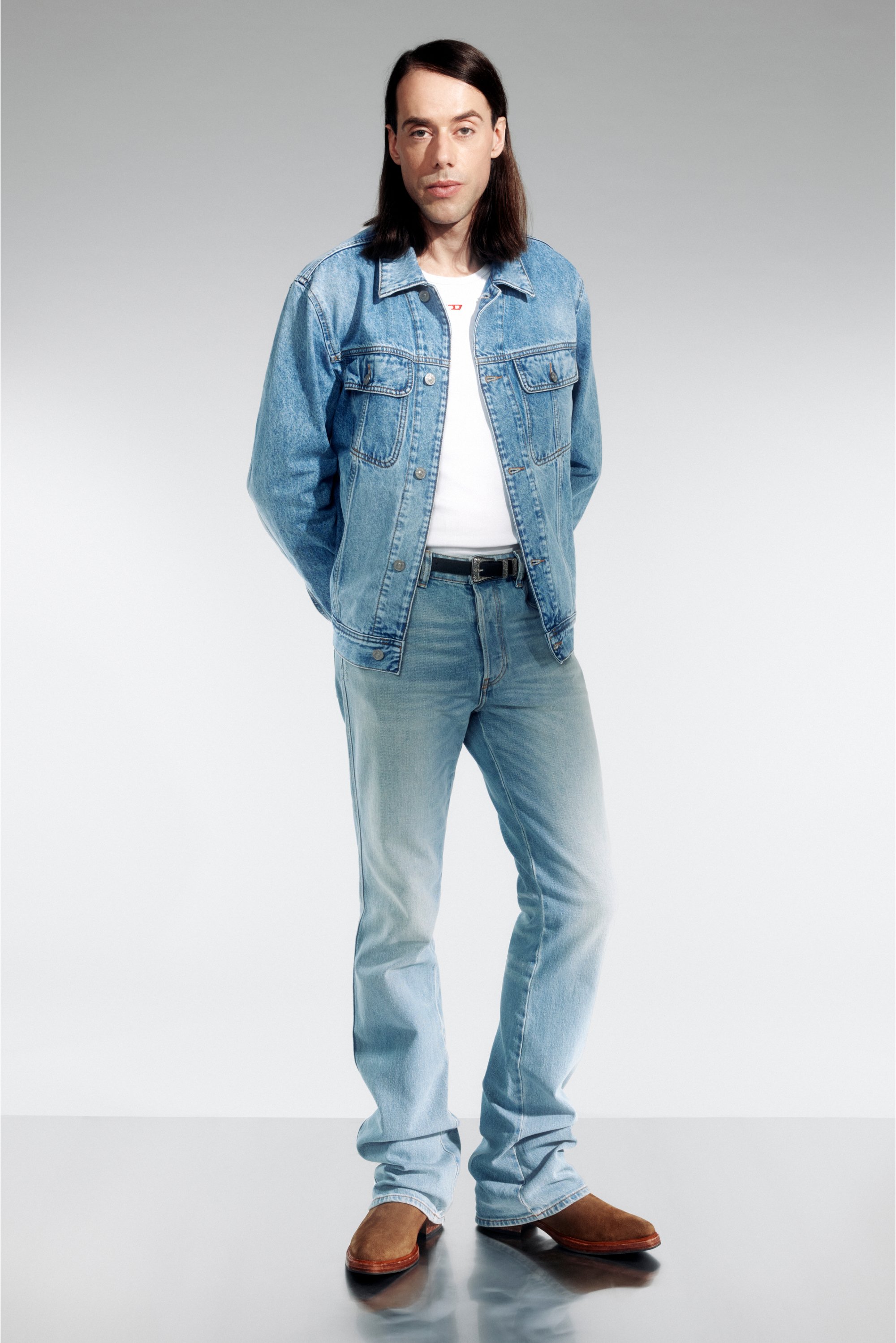 Man with long hair stands confidently in a denim jacket over a white t-shirt, paired with light jeans and brown boots, against a gray backdrop.