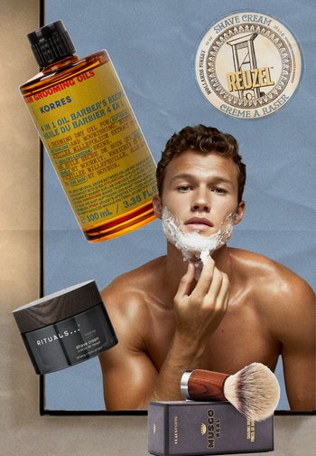 Jeune homme appliquant de la crème à raser avec un pinceau à raser, entouré de flacons d'huile de soin et de contenants de crème à raser sur un fond bleu.