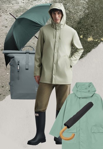 Man in een beige regenjas met kap, bruine broek, zwarte regenlaarzen, staand naast een grijze rugzak, zwarte paraplu en groene regenjas met houten handvat paraplu.