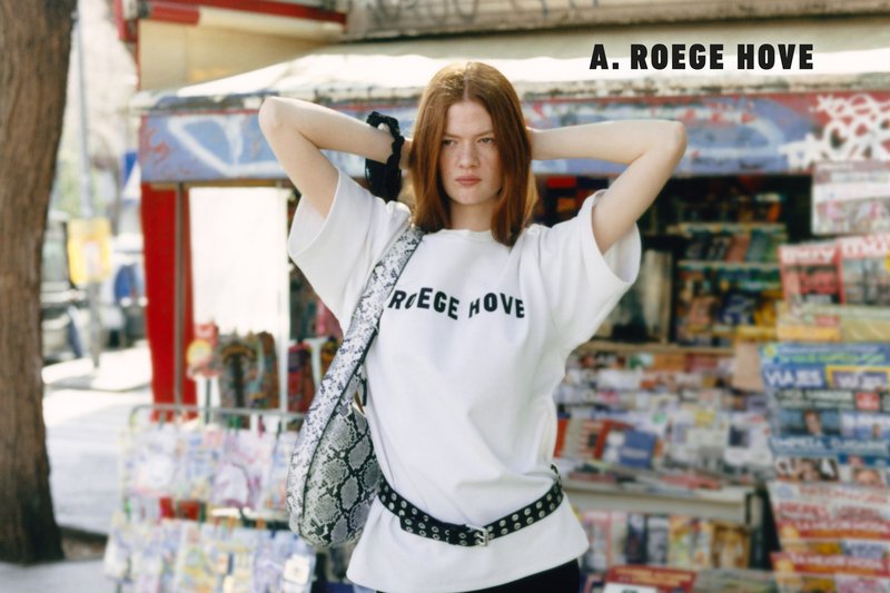 Junge Frau mit roten Haaren, die ein weißes A. Roege Hove Shirt und eine gemusterte Tasche trägt, steht mit den Händen hinter dem Kopf an einem Zeitungskiosk.