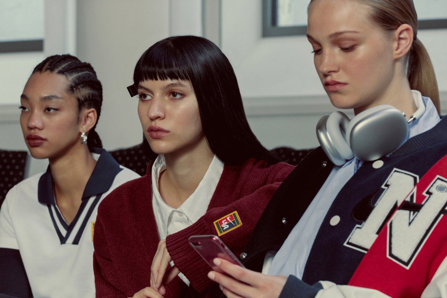 Tre giovani donne siedono affiancate, vestite con abiti casual e alla moda, mentre una tiene un telefono e un'altra indossa delle cuffie.