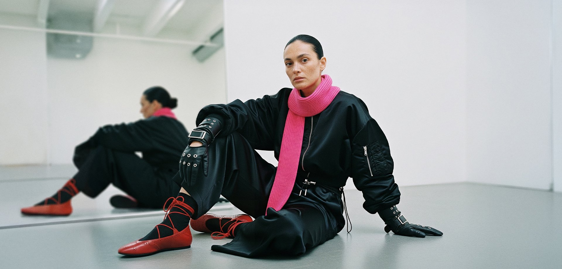 Black oversized outfit with a pink scarf, studded gloves, and red footwear with crisscrossed straps. Neutral background.