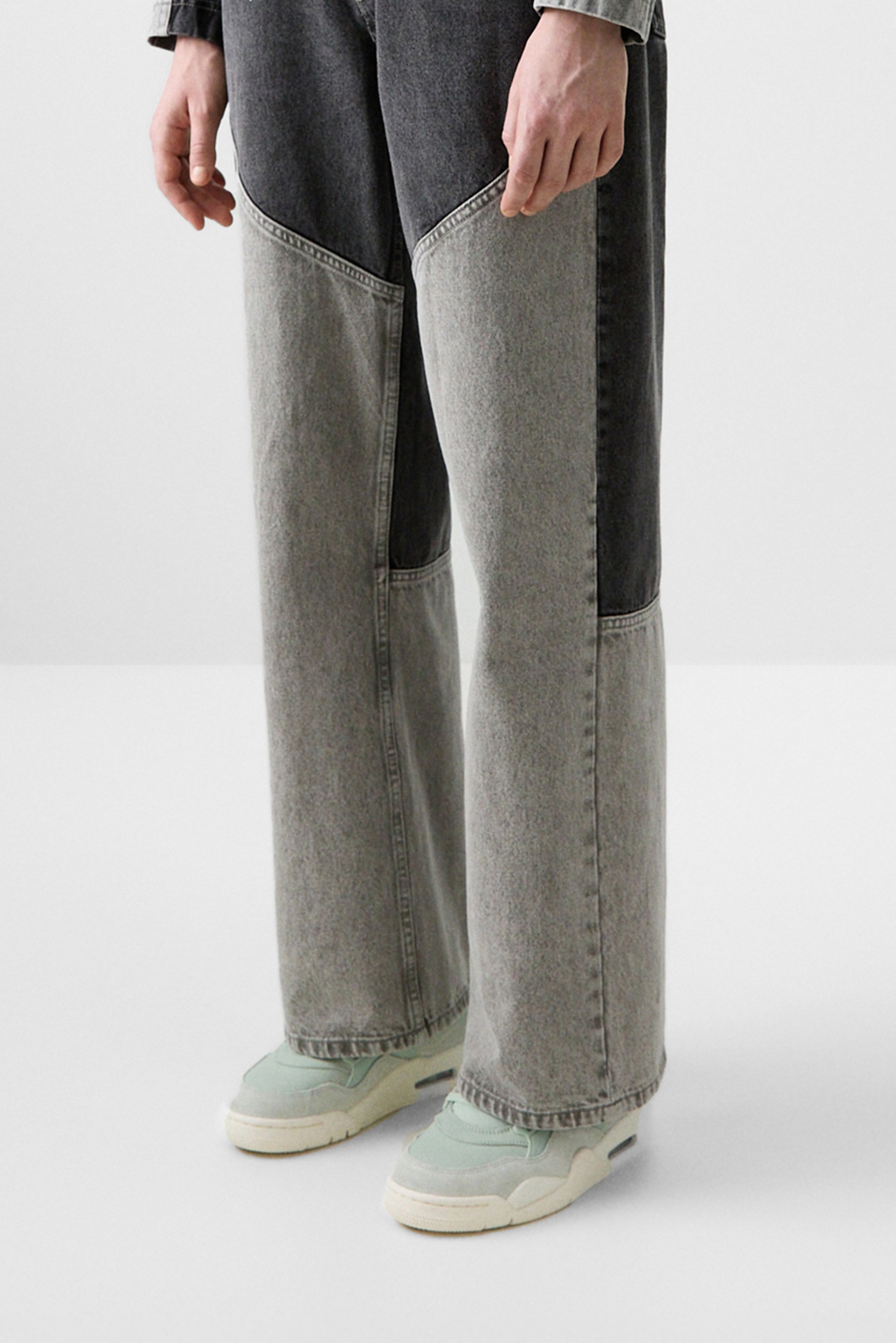 Una persona sta in piedi indossando pantaloni di jeans oversize unici con pannelli neri e grigi a contrasto, abbinati a sneakers verde chiaro.