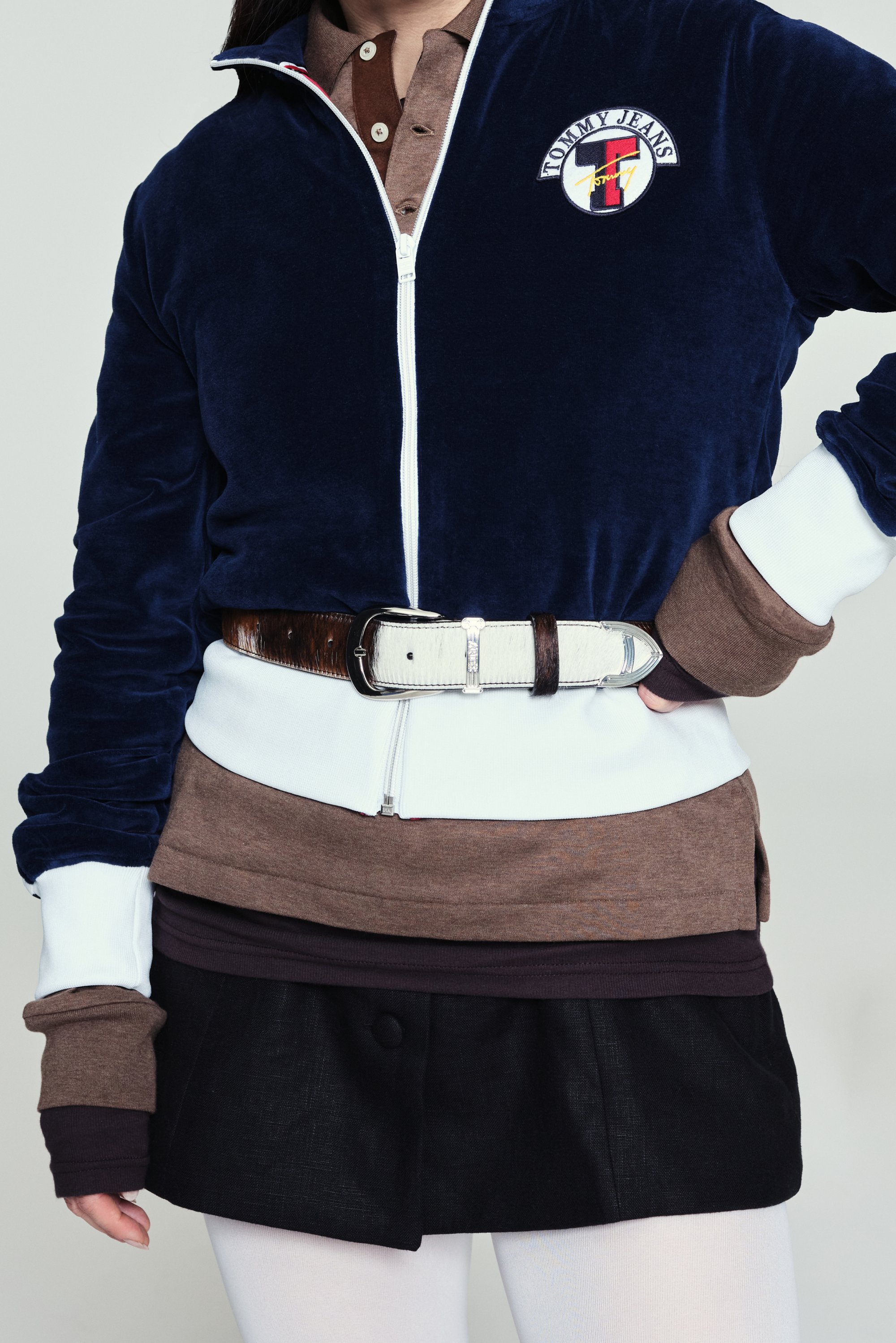 Giacca zip-up in velluto blu navy con accenti bianchi e marroni, indossata sopra top marroni e viola scuro, abbinata a una gonna nera e una cintura.