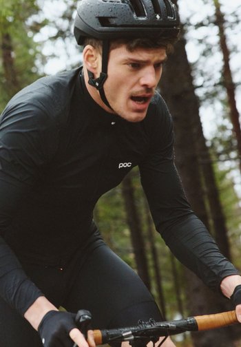 Syklist iført svart hjelm og klær sykler på en sykkel med brune styrehåndtak gjennom et skogsområde.