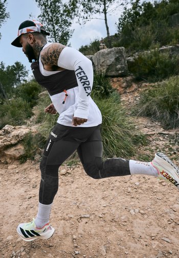 Man with tattoos runs on rocky trail wearing white Terrex arm sleeves, black leggings, white socks, Adidas trainers, and a black and white cap.